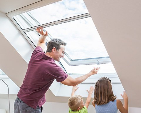 Ein Vater schaut mit seinen Kindern aus einem Dachfenster mit Fliegengitter-Rollo